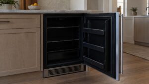 A dark, unlit built-in undercounter refrigerator with its door open, showing no interior light or display activity, kitchen cabinetry around it