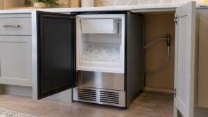 An under-counter ice machine installed in cabinetry with its door open and a thin water supply line visible running toward the cabinet wall