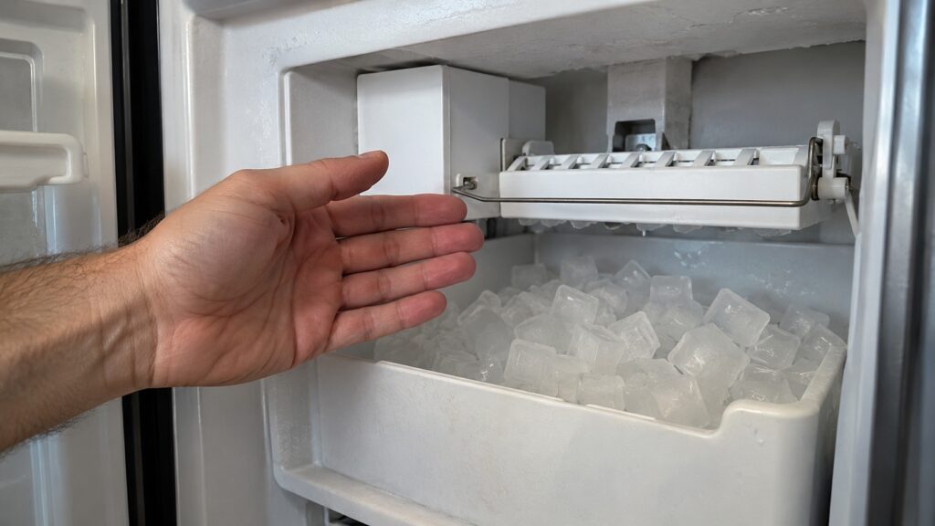 A hand beside an open ice bin showing a thin metal arm across the top of the ice compartment